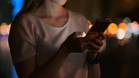 Close-up-of-a-mobile-phone-in-the-hands-of-a-girl-presses-her-fingers-on-the-screen-in-the-night-city-on-the-background-of-a-beautiful-bokeh.-Young-businessman-girl